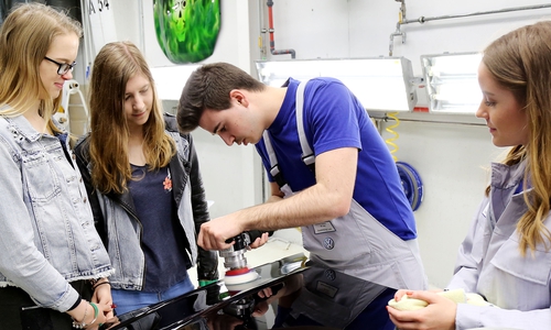 Sarah Bischoff (16, links) und Anna-Lena Bänsch (15) von der Realschule Wolfsburg-Vorsfelde schauen den Auszubildenden zu Fahrzeuglackierern, Sandro Lessi (22) und Merle Nieß (20, ganz rechts) beim Polieren einer Motorhaube zu. Fotos: Volkswagen/ Matthias Leitzke