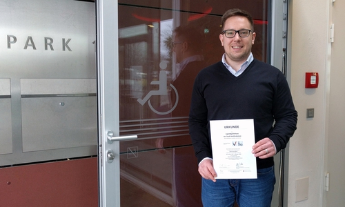 Betriebsleiter Benjamin Höfener mit der Urkunde „Reisen für alle“ vor dem Zugang für Rollstuhlfahrer, der neu gekennzeichnet wurde und nun sensorgesteuert ist. Foto: Stadt Wolfenbüttel