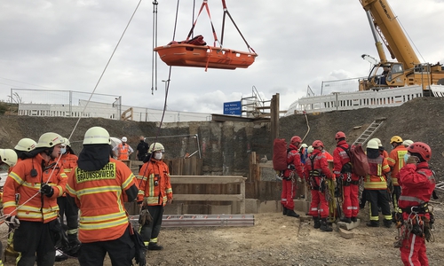 Die Feuerwehr bringt eine Rettungstrage, befestigt an einem Drehleiterfahrzeug in Position. Im Hintergrund die mehrere Meter tiefe Baugrube. 