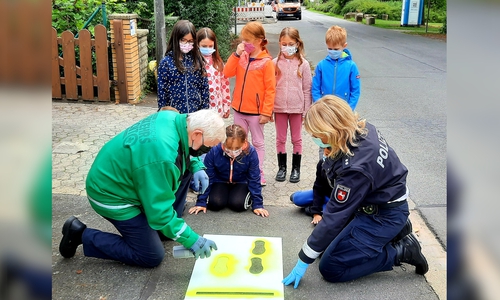 Polizei und Verkehrswacht beim aufsprühen der gelben Füße auf dem Bürgersteig. Sie bringen die Schülerinnen und Schüler dorthin, wo sie sicher die Straße überqueren können.
