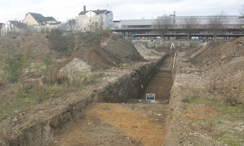 Einer der archäologischen Suchschnitte mit dem deutlichem Geländeabfall der ehemaligen Kiesgrube nach Norden. Im Hintergrund die Rückseite des Peiner Bahnhofs. 