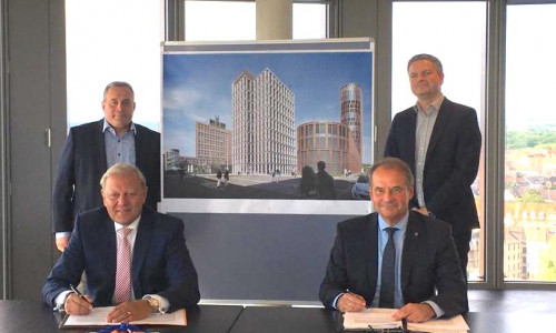 Jürgen Brinkmann (vorne links) und Ulrich Markurth (vorne rechts) bei der Vertragsunterzeichnung. Im Hintergrund sind Klaus Gattermann (links) und Carsten Beddig (rechts) zu sehen. Zwischen ihnen ist das geplante Gebäude zu erkennen.
