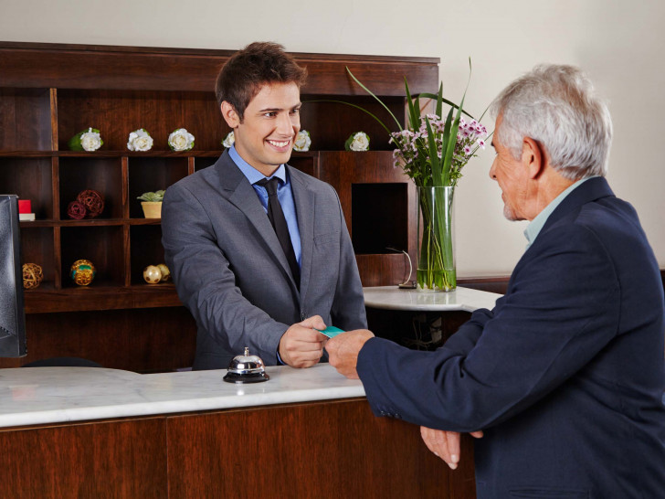 Ein junger Rezeptionist übergibt einem älteren Gast eine Zimmerkarte. Foto: Bundesagentur für Arbeit