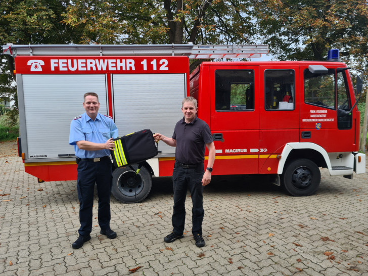 Schornsteinfegermeister Sven Holze (rechts) überreicht den Schornsteinfegerrucksack an einen Vertreter der Feuerwehr. 