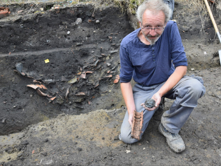 Grabungsleiter Bernhard Thiemann zeigt ein Kachelbruchstück mit Löwenkopf und eine Modell zur Herstellung von Ofenkacheln. Im Hintergrund ist eine Grube zu sehen, in der viel Keramik geborgen wurde.
