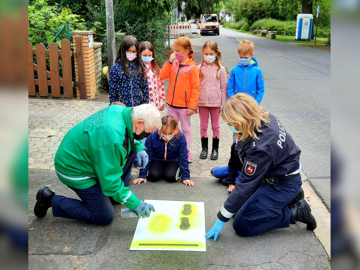 Polizei und Verkehrswacht beim aufsprühen der gelben Füße auf dem Bürgersteig. Sie bringen die Schülerinnen und Schüler dorthin, wo sie sicher die Straße überqueren können.