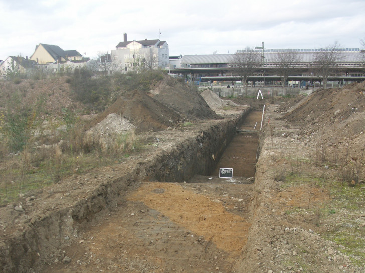 Einer der archäologischen Suchschnitte mit dem deutlichem Geländeabfall der ehemaligen Kiesgrube nach Norden. Im Hintergrund die Rückseite des Peiner Bahnhofs. 