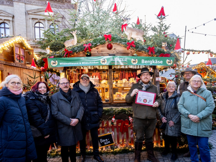 Der schönste Stand auf dem Braunschweiger Weihnachtsmarkt 2025 steht fest: „Harzer Spezialitäten“ bekommt in diesem Jahr die Auszeichnung überreicht: (v. l. n. r.) Cornelia Götz, Carolin Illmer, Karsten Ziaja, Olaf Jaeschke, Standbetreiber Franko Almstadt, Ulrike Neumann, Thomas Bronswyk und Bezirksbürgermeisterin Jutta Plinke.